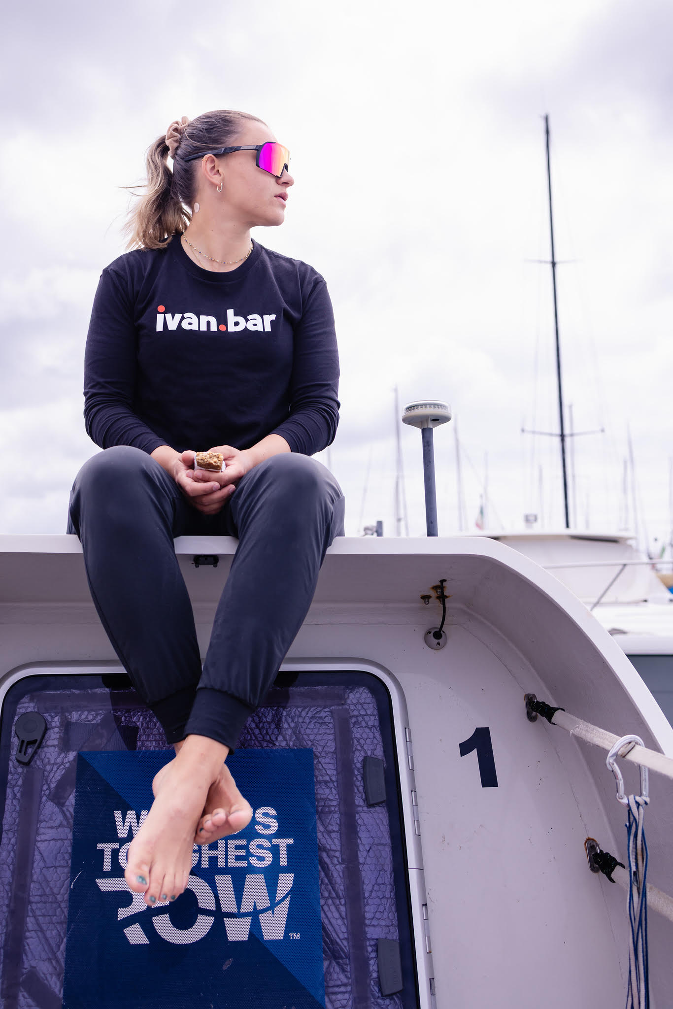A Sea Birds rower in an ivan.bar shirt sits atop their World's Toughest Row ocean rowing boat at the marina